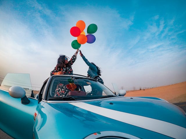 People driving with the car roof down. They are holding balloons.