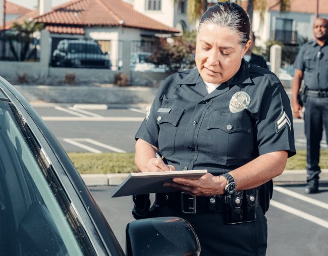 A police officer standing next to a car while writing something on a clip board.